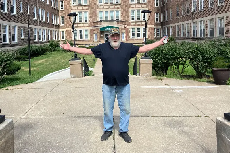 Jack West, a white man, with a white beard stands in front of a brown-bricked apartment building with his arms wide open.